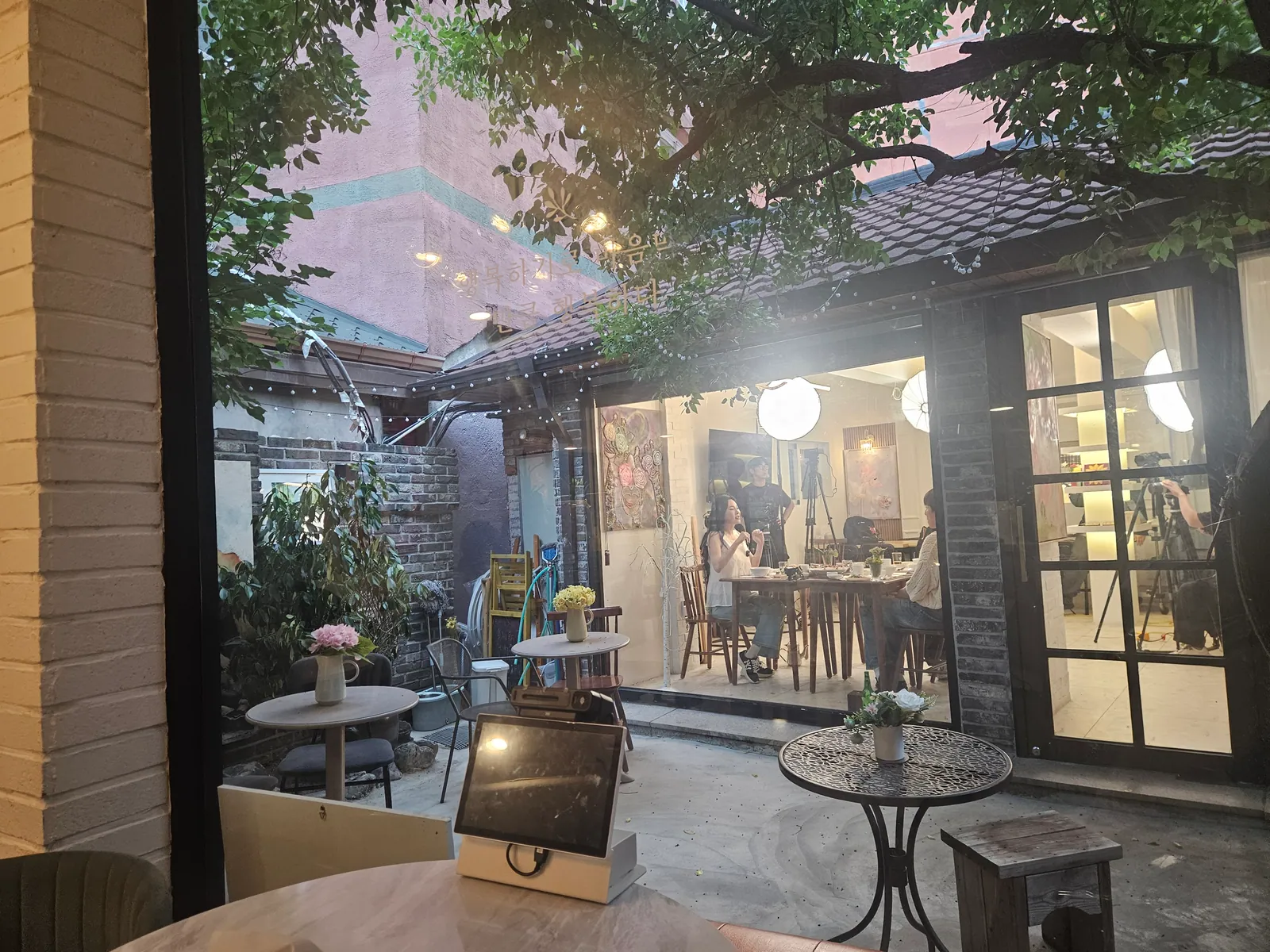 夕暮れの中庭。軒に吊るした明かりが灯り、窓の向こうのホールが明るい。