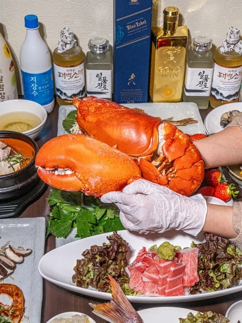 저녁 상차림 세로 컷. 상 뒤로 전통주와 위스키가 늘어서 있고, 앞에는 랍스터와 회, 뚝배기가 놓여 있다.