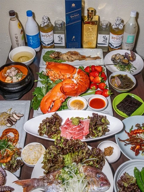 저녁 상차림 세로 컷. 상 뒤로 전통주와 위스키가 늘어서 있고, 앞에는 랍스터와 회, 뚝배기가 놓여 있다.